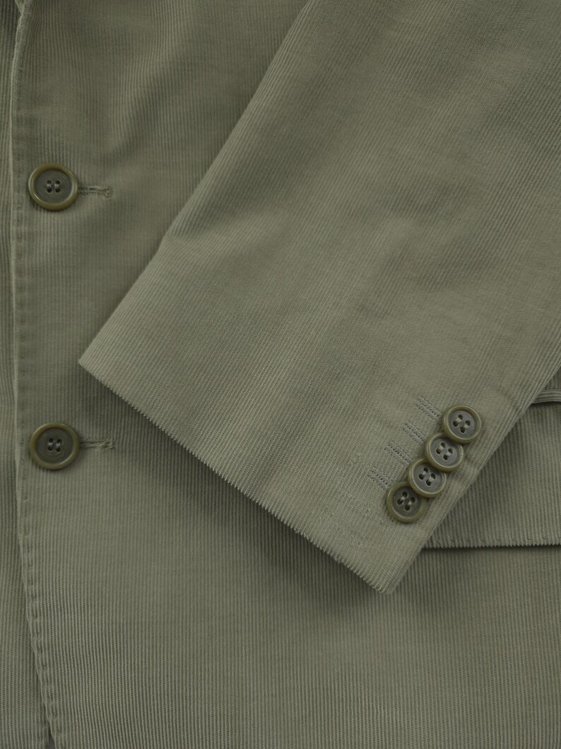 May include: Close-up of a sage green corduroy blazer. The blazer features three buttons on the sleeve cuff and two on the front. The fabric has a ribbed texture.