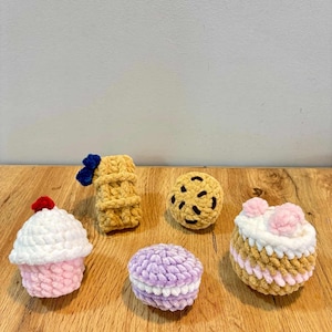 May include: A collection of crocheted food items, including a cupcake with a red cherry, a waffle with a blue bow, a chocolate chip cookie, a macaron, and a cake with pink flowers. The items are arranged on a wooden surface.