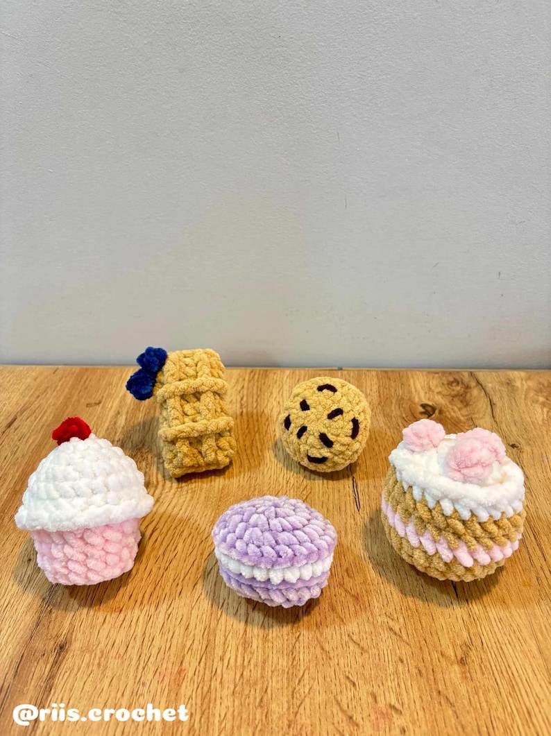 May include: A collection of crocheted food items, including a cupcake with a red cherry, a waffle with a blue bow, a chocolate chip cookie, a macaron, and a cake with pink flowers. The items are arranged on a wooden surface.