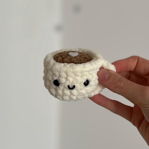May include: A crocheted coffee cup with a smiling face, black eyes, and a curved mouth. The cup is off-white with a brown interior and a white heart detail. The handle is visible on the right side.