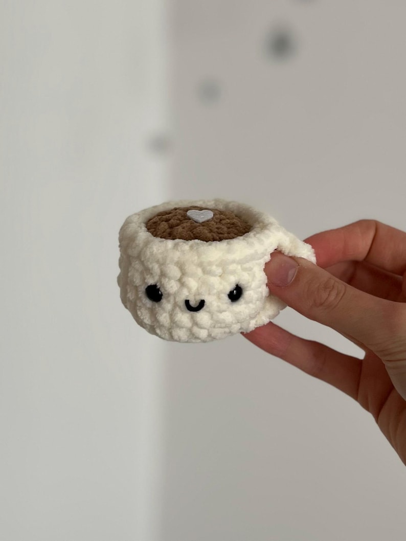 May include: A crocheted coffee cup with a smiling face, black eyes, and a curved mouth. The cup is off-white with a brown interior and a white heart detail. The handle is visible on the right side.
