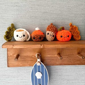 May include: A wooden shelf displays crocheted autumn-themed items: a coffee cup, a cupcake, an acorn, and a pumpkin, all with smiling faces. Below, a blue and white striped decorative door with a seashell and hook hangs. The shelf has wooden pegs.