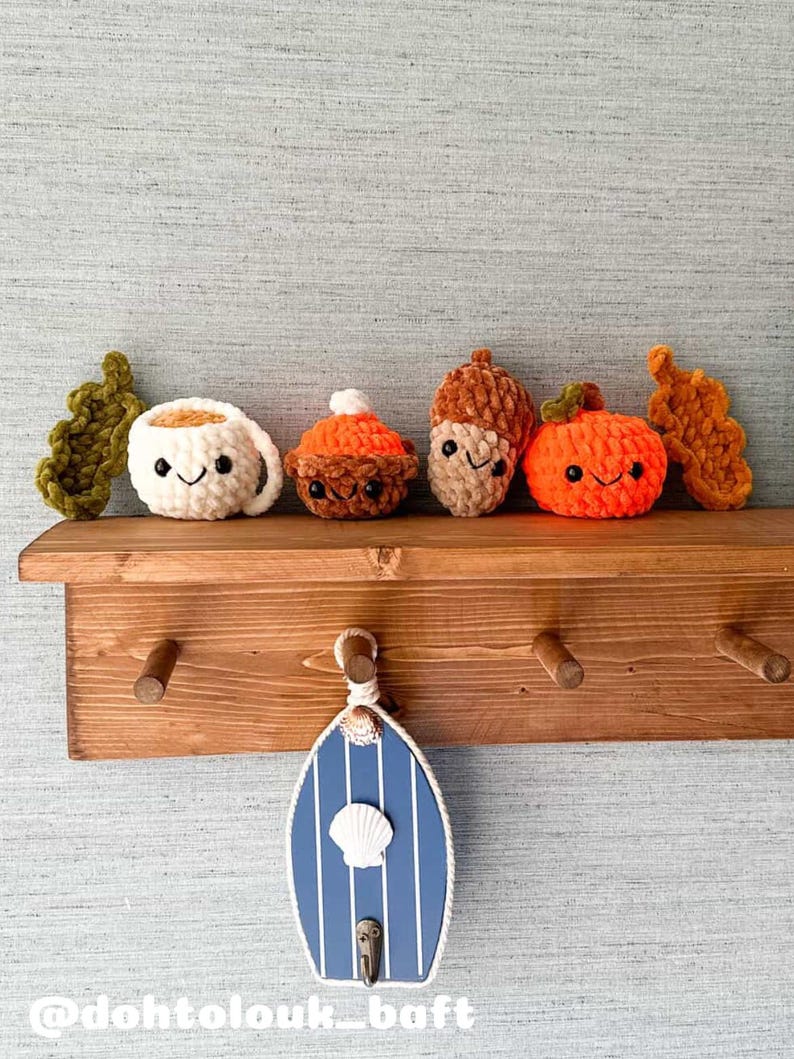 May include: A wooden shelf displays crocheted autumn-themed items: a coffee cup, a cupcake, an acorn, and a pumpkin, all with smiling faces. Below, a blue and white striped decorative door with a seashell and hook hangs. The shelf has wooden pegs.