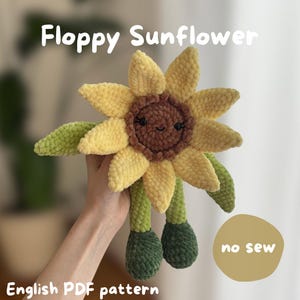 Könnte beinhalten: Ein gehäkeltes Sonnenblumen-Spielzeug mit einem lächelnden Gesicht, gelben Blütenblättern und einer braunen Mitte. Es hat grüne Blätter und Beine. Der Text "Floppy Sunflower" und "English PDF pattern" sind sichtbar.