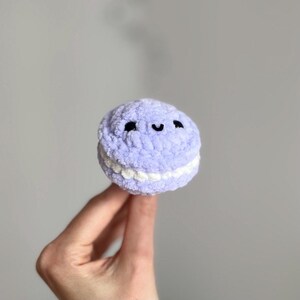 May include: A lavender crocheted macaron with a white filling and a smiling face. The plush toy has black eyes and a curved mouth. The soft, round dessert is held in a hand against a gray background.
