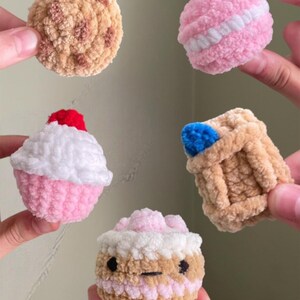 May include: A collection of handmade crochet food items, including a cookie, a pink macaron, a cupcake with a red cherry, a small basket with a blue item, and a cupcake with a face. The items are in various shades of pink, brown, and white. The text "@bunnyhopcrochets" is at the bottom.