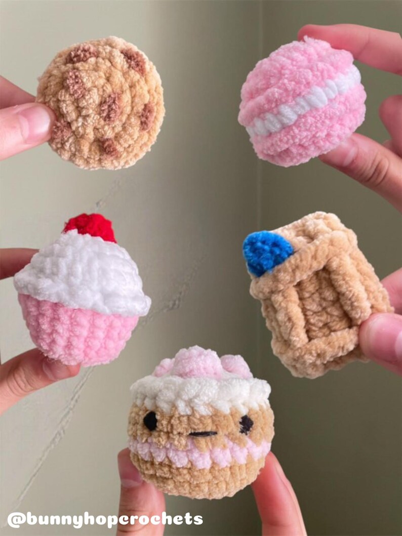 May include: A collection of handmade crochet food items, including a cookie, a pink macaron, a cupcake with a red cherry, a small basket with a blue item, and a cupcake with a face. The items are in various shades of pink, brown, and white. The text "@bunnyhopcrochets" is at the bottom.