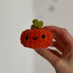May include: A small, orange crocheted pumpkin with a smiling face and a green stem. The pumpkin is held in a hand, showcasing its soft, textured appearance. This handmade item is perfect for autumn decor.