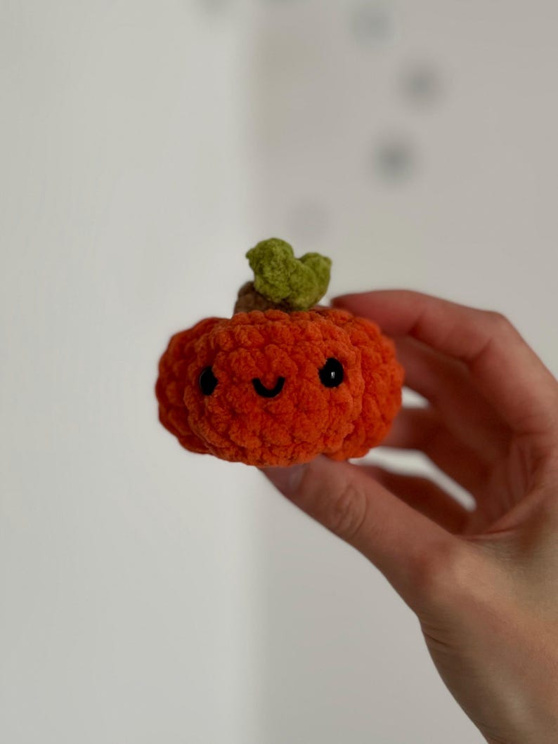 May include: A small, orange crocheted pumpkin with a smiling face and a green stem. The pumpkin is held in a hand, showcasing its soft, textured appearance. This handmade item is perfect for autumn decor.