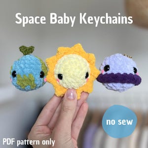Peut inclure: Porte-clés crochetés sur le thème de l'espace. Les porte-clés comprennent une Terre bleue et verte, un soleil jaune et une planète violette avec un anneau. Le texte "Space Baby Keychains" et "PDF pattern only" sont visibles.