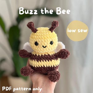 Può includere: Un peluche a forma di ape all'uncinetto, con corpo giallo, strisce marroni e piccole ali. Il testo "Buzz the Bee" è in alto, con "low sew" e "PDF pattern only" visibili. L'ape ha una faccia sorridente.