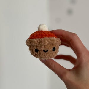 May include: A crocheted pumpkin pie with a smiling face and a white dollop of whipped cream on top. The pie has a brown crust, an orange filling, and black eyes. The handmade pie is held in a hand.