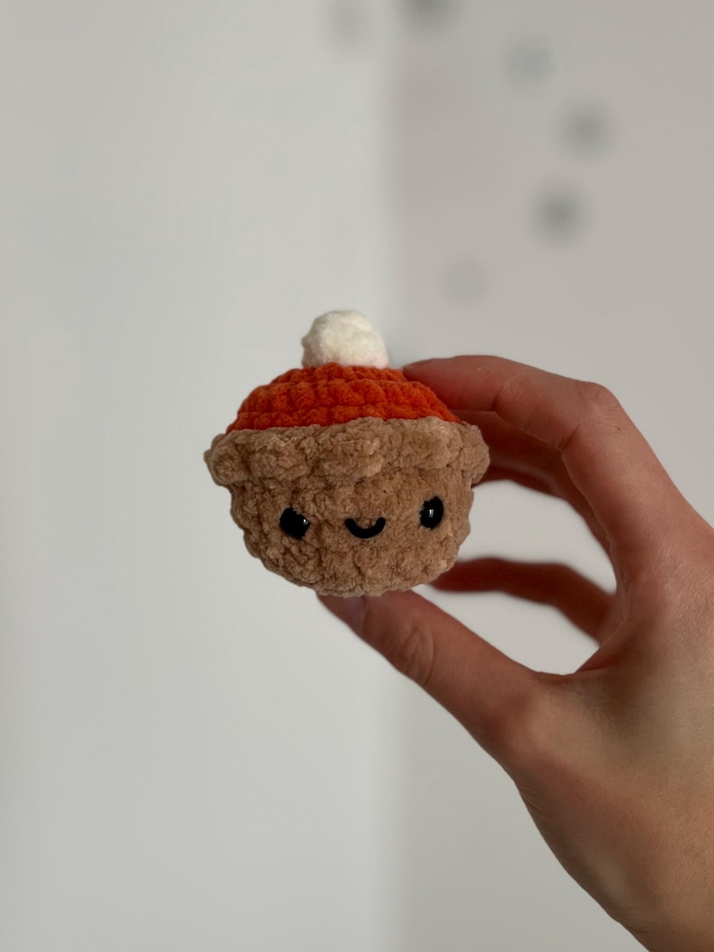 May include: A crocheted pumpkin pie with a smiling face and a white dollop of whipped cream on top. The pie has a brown crust, an orange filling, and black eyes. The handmade pie is held in a hand.