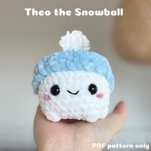 Può includere: Un giocattolo di peluche a forma di palla di neve bianca all'uncinetto, che indossa un cappello blu all'uncinetto con un pon pon bianco. Il giocattolo ha occhi neri, un blush rosa e una bocca sorridente. Il testo "Theo the Snowball" è sopra il giocattolo e "PDF pattern only" è sotto il giocattolo.