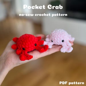 Puede incluir: Dos juguetes de cangrejo de ganchillo, uno rojo y otro rosa, se sostienen en una mano. El cangrejo rojo está a la izquierda y el cangrejo rosa a la derecha. El texto "Pocket Crab" y "no-sew crochet pattern" están en la parte superior. "PDF pattern" está en la parte inferior.