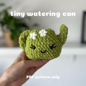 Könnte beinhalten: Eine grüne gehäkelte Gießkanne mit weißen Blumenakzenten und einem lächelnden Gesicht. Der Text "tiny watering can" und "PDF pattern only" ist im Bild sichtbar.