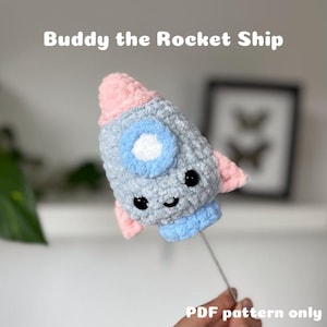 May include: A crocheted rocket ship toy with a pink and blue color scheme. The rocket has a smiling face with black eyes and a white mouth. The text "Buddy the Rocket Ship" is at the top of the image. The text "PDF pattern only" is at the bottom of the image.