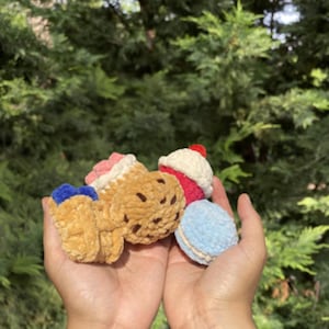 May include: A collection of handmade crochet food items, including a cupcake, ice cream cone, cookie, and macaron. The items are in various colors, such as pink, red, blue, and brown. The Instagram handle @strawberrycrochets is visible at the bottom.