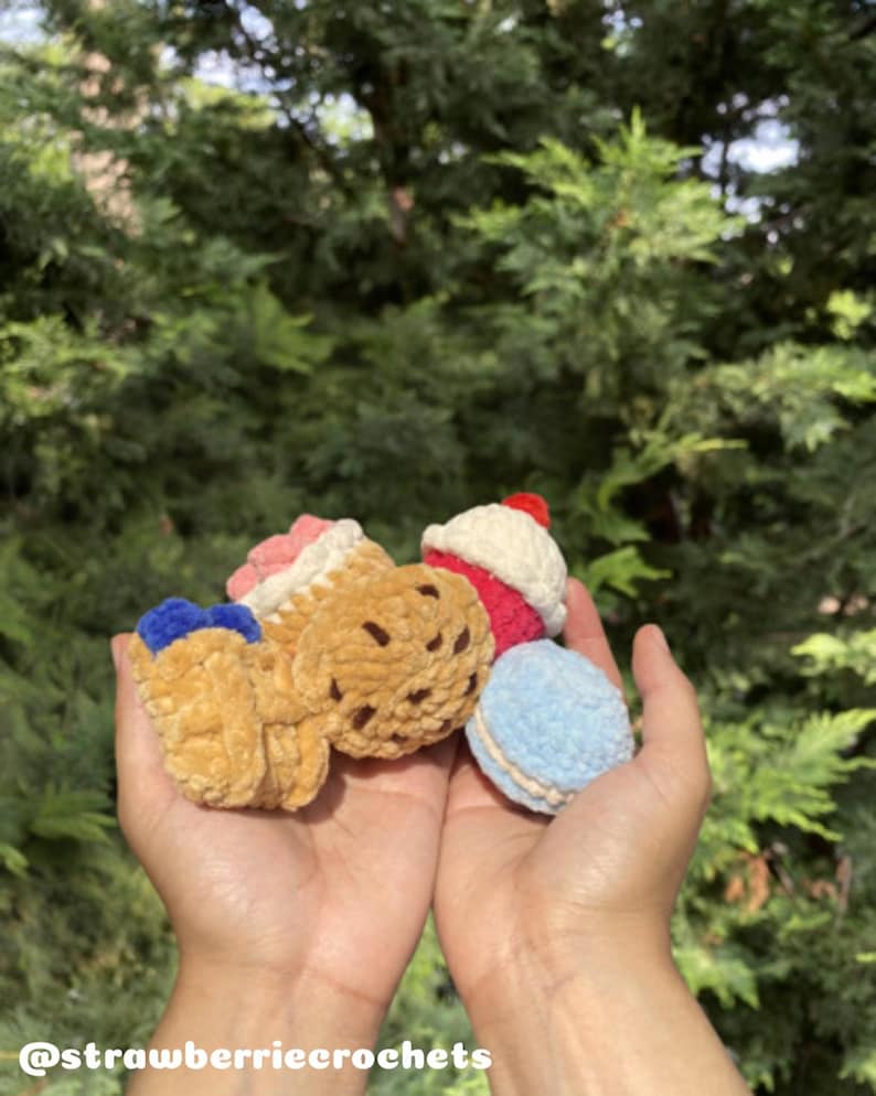 May include: A collection of handmade crochet food items, including a cupcake, ice cream cone, cookie, and macaron. The items are in various colors, such as pink, red, blue, and brown. The Instagram handle @strawberrycrochets is visible at the bottom.