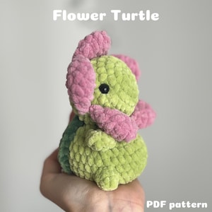 Puede incluir: Un juguete de tortuga de peluche hecho a mano con un diseño de flor. La tortuga es principalmente verde claro con pétalos de flores rosas y un caparazón verde oscuro. El texto "Flower Turtle" y "PDF pattern" son visibles.