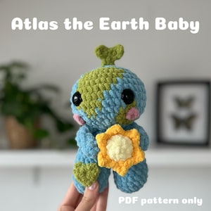 Op de afbeelding: Haakpatroon voor een blauw en groen aarde-vormig amigurumi-speelgoed dat een gele zon vasthoudt. Het patroon heet "Atlas the Earth Baby" en is beschikbaar als PDF-download.