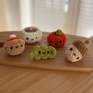 May include: A selection of crocheted food items, including a pumpkin, acorn, cinnamon roll, and a bean pod, each with a smiling face. The items are in shades of orange, brown, green, and cream, displayed on a wooden chopping board.