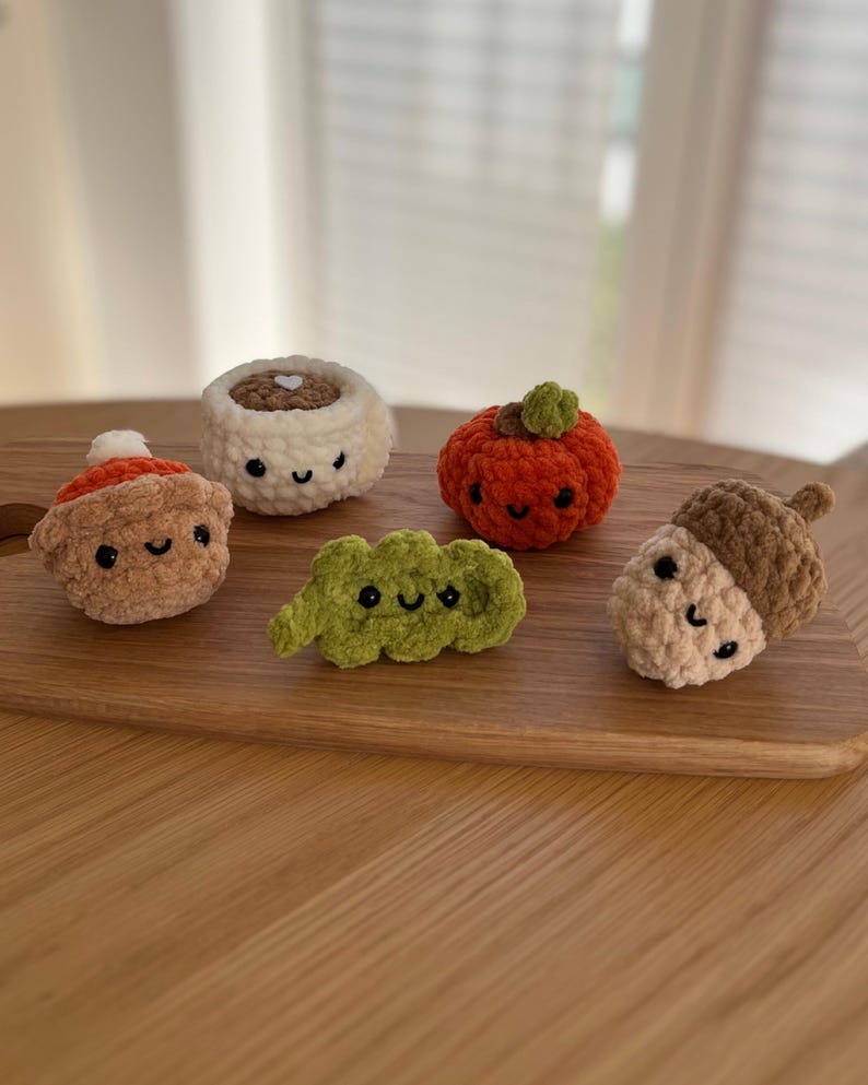 May include: A selection of crocheted food items, including a pumpkin, acorn, cinnamon roll, and a bean pod, each with a smiling face. The items are in shades of orange, brown, green, and cream, displayed on a wooden chopping board.
