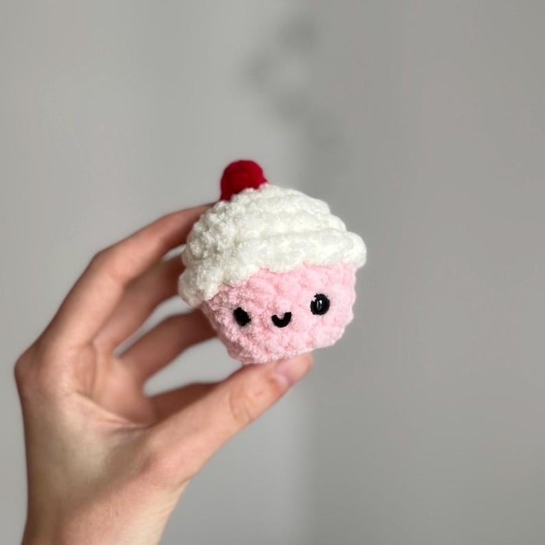 May include: A handmade, plush cupcake with a pink base and white frosting. The cupcake has a smiling face with black eyes and a curved mouth. A small red cherry tops the frosting. The cupcake is held in a hand.