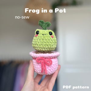 May include: A crochet pattern for a frog in a pot. The frog is green with a pink pot and a pink bow. The pattern is no-sew and is available as a PDF download.