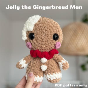 Op de afbeelding: Een gehaakt knuffel van een gingerbread man met witte glazuurdetails, een rode strik en zwarte ogen. Het speelgoed wordt in een hand gehouden en de tekst "Jolly the Gingerbread Man" en "PDF pattern only" is zichtbaar.