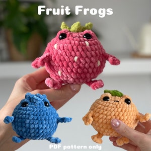 Op de afbeelding: Drie gehaakte pluche speelgoedjes in de vorm van kikkers, elk met een ander fruitontwerp. De eerste kikker is roze en heeft de vorm van een aardbei, de tweede is blauw en de derde is oranje. De speelgoedjes staan allemaal op een wit oppervlak. De tekst "Fruit Frogs" staat bovenaan de afbeelding in het zwart. De tekst "PDF pattern only" staat onderaan de afbeelding in het zwart.