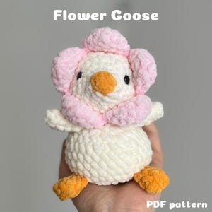 Könnte beinhalten: Ein handgefertigtes gehäkeltes Plüschtier in Form einer Gans mit Blumendesign. Die Gans ist weiß mit einem rosa Blütenkopf und -kragen sowie einem orangefarbenen Schnabel und Füßen. Der Text "Flower Goose" steht oben und "PDF pattern" unten.