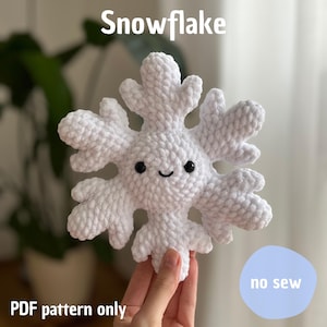 Könnte beinhalten: Ein weißer, gehäkelter Schneeflocken-Anhänger mit einem lächelnden Gesicht und schwarzen Augen. Der Text "Snowflake" steht oben, und "PDF pattern only" steht unten. Die Worte "no sew" stehen in einem blauen Kreis.
