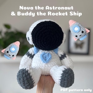 May include: A crochet pattern for a white astronaut with a black helmet and a blue heart, and a small gray and pink rocket ship. The pattern is called "Nova the Astronaut & Buddy the Rocket Ship" and is available as a PDF download.