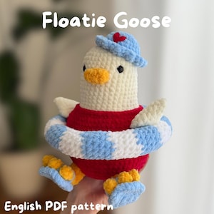 Könnte beinhalten: Ein gehäkeltes Plüschgans-Spielzeug namens Floatie Goose mit weißem Körper, orangefarbenem Schnabel und Füßen. Es trägt ein rotes Oberteil, einen blauen Hut mit rotem Herz und einen blau-weiß gestreiften Schwimmring. Text: Floatie Goose und English PDF pattern.