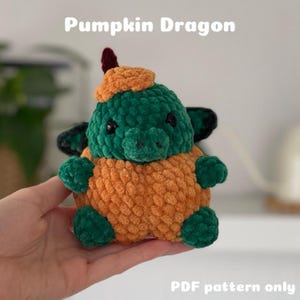 Könnte beinhalten: Ein gehäkeltes Kürbis-Drachen-Plüschtier aus grünem und orangefarbenem Garn. Der Drache trägt einen kleinen, spitzen Hut auf dem Kopf. Der Text "Pumpkin Dragon" befindet sich oben im Bild und "PDF pattern only" unten.