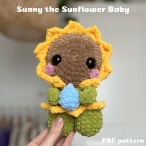 Op de afbeelding: Een gehaakt zonnebloem baby speelgoed met een bruin gezicht, gele bloemblaadjes, groene bladeren en een blauwe waterdruppel. Het speelgoed wordt vastgehouden door een hand. De tekst "Sunny the Sunflower Baby" en "PDF pattern" is zichtbaar in de afbeelding.