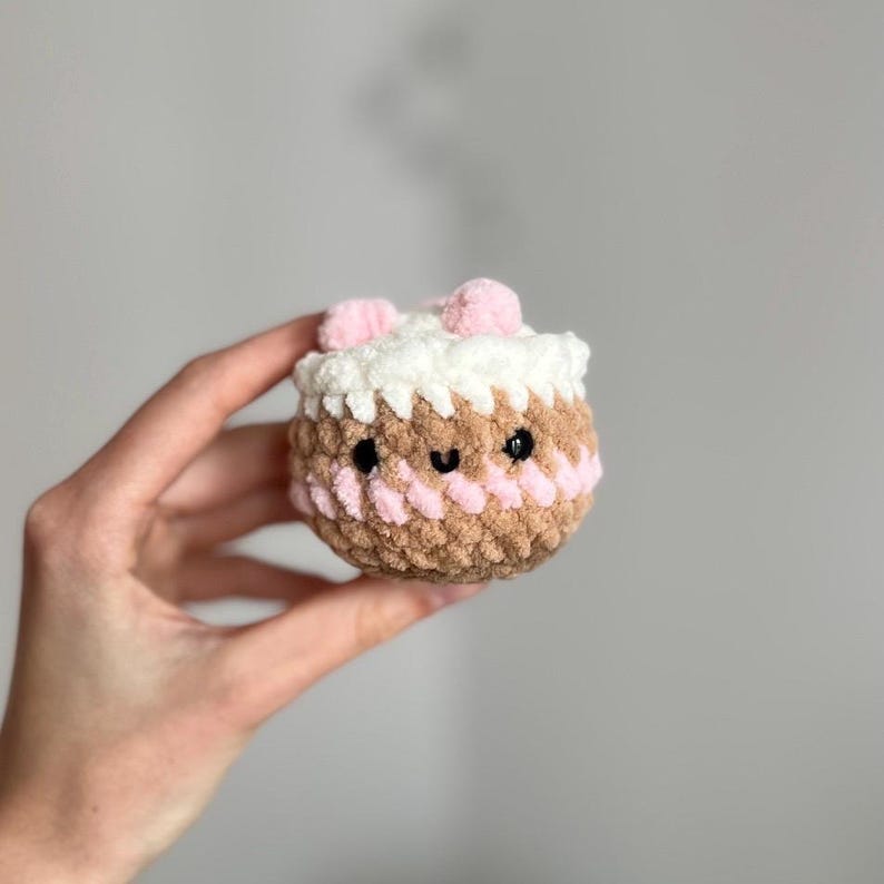 May include: A small, round, crocheted cake with a smiling face. The cake features a brown base, a pink stripe, white frosting, and pink pom-poms on top. Black eyes and a small black mouth complete the design.