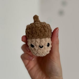 May include: A crocheted acorn toy with a smiling face. The top of the acorn is a dark brown, and the body is a light beige colour. The eyes are black, and the mouth is a curved black line. The toy is held in a hand.