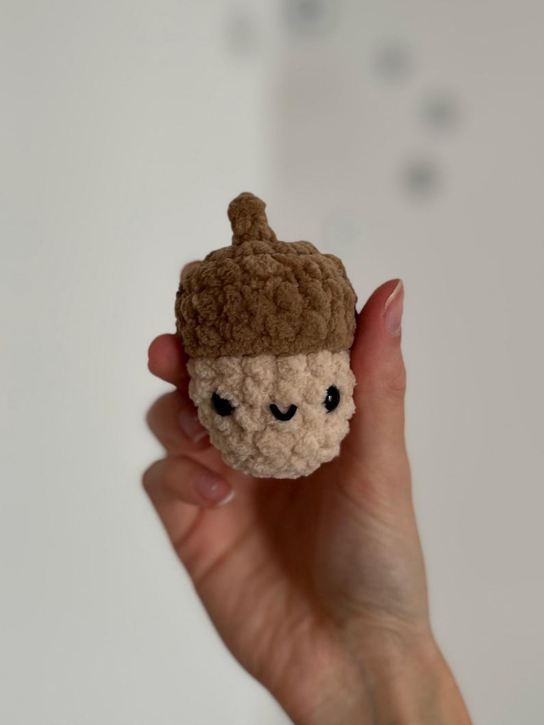 May include: A crocheted acorn toy with a smiling face. The top of the acorn is a dark brown, and the body is a light beige colour. The eyes are black, and the mouth is a curved black line. The toy is held in a hand.