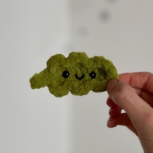 May include: A handmade, green crocheted cloud with black eyes and a smiling mouth. The cloud is held in a hand against a white background. The cloud is a whimsical, soft, and textured item.