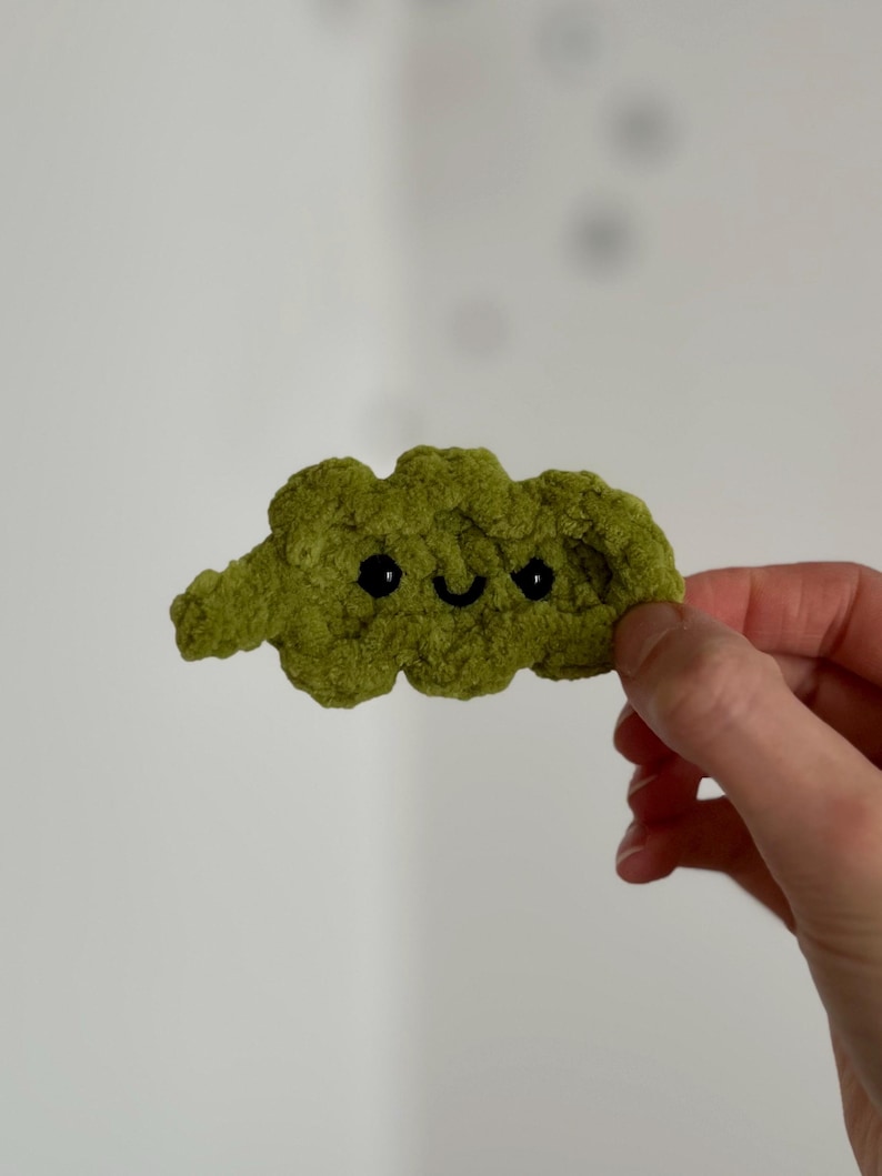 May include: A handmade, green crocheted cloud with black eyes and a smiling mouth. The cloud is held in a hand against a white background. The cloud is a whimsical, soft, and textured item.