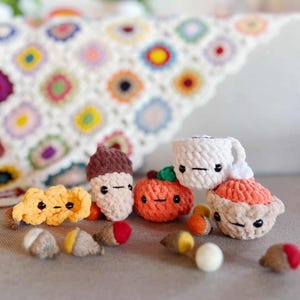 May include: A selection of crocheted autumn-themed items, such as an acorn, pumpkin, leaf, and coffee cup. The items are in autumnal colours of brown, orange, and yellow. A colourful, patterned blanket is in the background. The text "@thegoodknotstudio" is at the bottom.