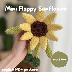 Peut inclure: Une fleur de tournesol en crochet avec des pétales jaunes, un centre marron foncé avec des yeux noirs et une bouche souriante. Elle possède des tiges et des feuilles vertes. Le texte indique : Mini Floppy Sunflower, no sew, English PDF pattern.