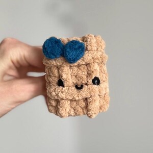 May include: A handmade, tan crochet cube with a smiling face and two blue pom-poms on top. The cube has black eyes and a curved mouth, giving it a friendly appearance. The textured yarn creates a soft, plush look.