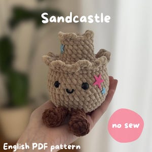 Puede incluir: Un juguete de castillo de arena tejido a ganchillo en hilo beige con una cara sonriente, ojos negros y acentos de estrella en rosa y azul. El castillo de arena tiene un borde con volantes y una torre. El texto "Sandcastle" está en la parte superior, y "no sew" está en un círculo rosa.