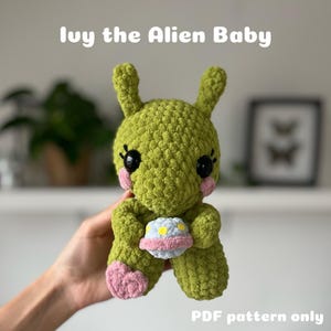 May include: A green crocheted alien baby toy holding a blue and yellow crocheted spaceship. The toy has pink cheeks and black eyes. The text "Ivy the Alien Baby" is above the toy and "PDF pattern only" is below the toy.