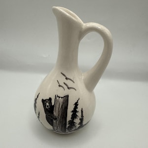 May include: A cream-colored ceramic pitcher with a narrow neck and a single handle. The body features a hand-painted black ink illustration of a bear peeking from behind a tree, surrounded by pine trees and birds flying in the sky above.