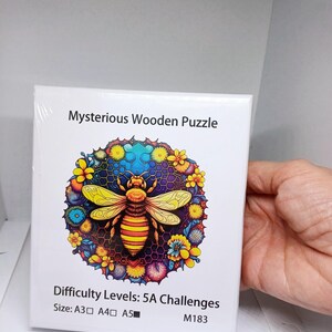 May include: A white box featuring a colourful image of a bee surrounded by flowers. The box is labelled "Mysterious Wooden Puzzle" and "Difficulty Levels: 5A Challenges". The box also includes the size options A3, A4, and A5, and the product code M183.