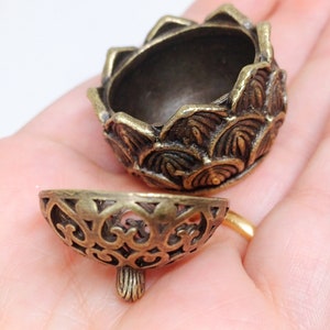 May include: An antique bronze-coloured decorative container with a detailed floral design. The container is in two parts, with a top and a separate, openwork bottom. The top has a petal-like design, while the bottom has an ornate, open filigree pattern.
